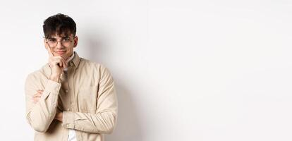 Cunning young man watching something interesting happen, lean face on arm and smiling devious, looking at camera intrigued, standing on white background photo
