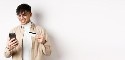 Online shopping. Happy young man using smartphone and plastic credit card, paying in internet, standing on white background photo