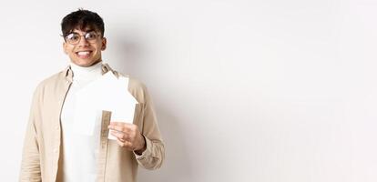 Real estate and mortgage concept. Cheerful young guy renting apartment, showing paper house cutout and smiling happy, buying property, white background photo