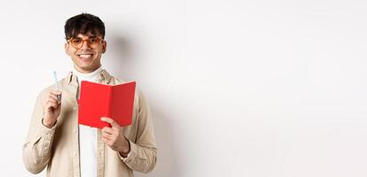 Happy person in glasses writing in journal, holding pen and diary, smiling at camera, plan a schedule, standing with planner on white background photo