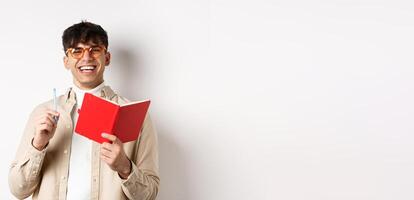 Cheerful young man in glasses laughing and taking notes, writing down in planner, holding pen and diary, standing on white background photo