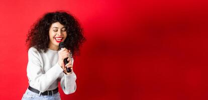 Hobbies and leisure concept. Beautiful woman singing in microphone and smiling, having fun at karaoke, standing on red background photo