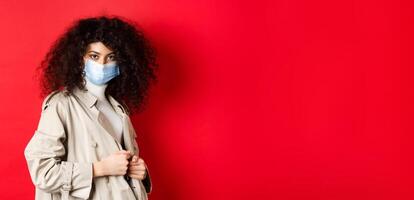 Covid-19, pandemic and quarantine concept. Sassy girl in trench coat and medical mask, put on trench coat for spring walk, red background photo
