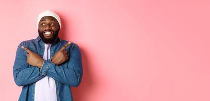 Excited young african-american man pointing sideways, showing two choices and making decision, smiling happy, standing over pink background photo