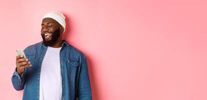 Technology and online shopping concept. Happy Black bearded man reading message and smiling, using smartphone against pink background photo