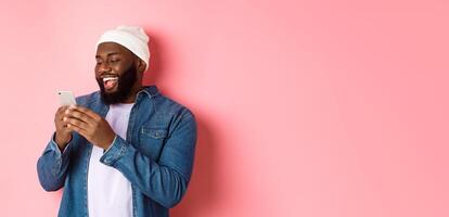 Technology and online shopping concept. Happy Black bearded man reading message and smiling, using smartphone against pink background photo