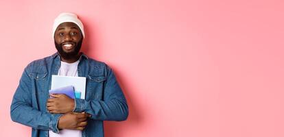 Image of adult Black man holding notebooks and smiling, studying at courses, standing over pink background photo