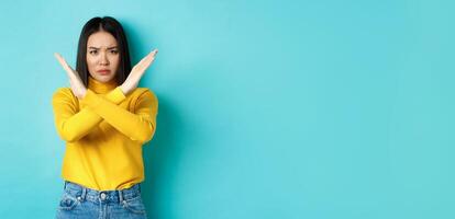 Serious and assertive korean woman showing cross stop gesture, frowning and telling no, prohibit action, disapprove something bad, standing over blue background photo