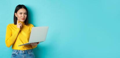 Serious looking asian woman working on laptop and thinking, frowning at camera, solving problem at work, standing over blue background photo