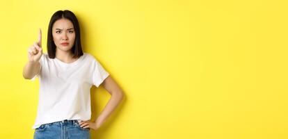 Confident and serious woman tell no, showing extended finger to stop and prohibit something bad, frowning and looking at camera self-assured, standing over yellow background photo