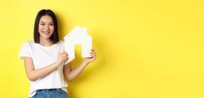 Real estate concept. Smiling beautiful woman showing paper house cutout and looking at camera, buying property, standing over yellow background photo