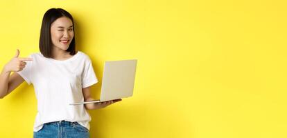 E-commerce and shopping concept. Good-looking asian girl winking at camera, smiling and pointing finger a laptop, showing something, standing over yellow background photo