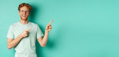 Handsome caucasian man with ginger hair, wearing glasses and t-shirt, pointing fingers right and smiling joyful, showing advertisement, standing over turquoise background photo