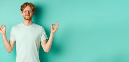 Calm and relaxed redhead man in glasses smiling, holding hands spread sideways with om mudra gesture, meditating peaceful, standing over mint background photo