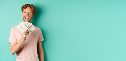 Happy redhead man in t-shirt showing money in dollars and smiling, making smug faces after winning cash, standing over turquoise background photo