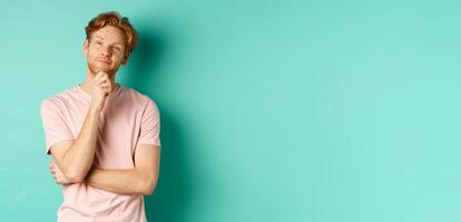 Pensive handsome man with red hair and beard looking at upper left corner, making choice and looking thoughtful, standing in t-shirt over mint background photo