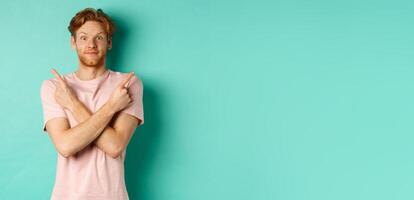 Attractive indecisive man in t-shirt cross hands and pointing sideways, showing two choices, need help with decision, standing over turquoise background photo