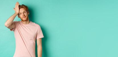 Annoyed and bothered redhead male model showing facepalm gesture, standing over mint background photo
