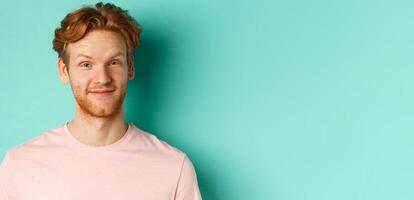 Close up of redhead bearded man looking pleased, nod in approval and smiling, standing in pink t-shirt against turquoise background photo