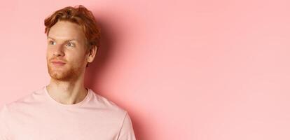 Handsome european male model with red hair and beard, turn head and looking pleased at copy space on left side, standing over pink background photo