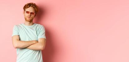Tourism and vacation concept. Sassy redhead guy in sunglasses, cross arms on chest and looking serious at camera, standing over pink background photo