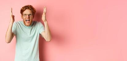 Angry redhead guy in glasses shouting, frowning and shaking hands with frustrated and outraged face, standing over pink background photo