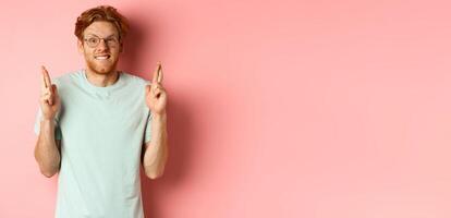 Worried redhead man waiting for results, expecting something with fingers crossed, biting finger and looking at something risky, standing over pink background photo