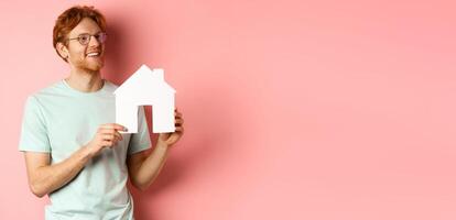 Real estate. Happy redhead man dreaming of buying property, looking right and showing paper house cutout, standing over pink background photo