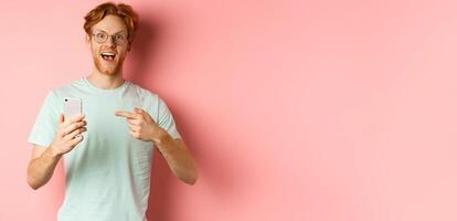Cheerful guy talking about internet promo, smiling amazed and pointing finger at smartphone, standing over pink background photo
