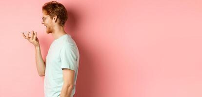 Profile portrait young man with red hair, smiling pleased while record voice message on smartphone, talking to virtual assistant, standing over pink background photo
