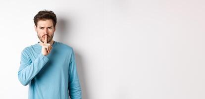 Angry man shushing at camera and telling to be quiet, frowning upset, standing over white background with finger on lips, taboo gesture photo