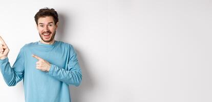 Happy caucasian man in casual blue sweatshirt, pointing fingers at upper left corner, showing link or logo on white background, smiling at camera photo