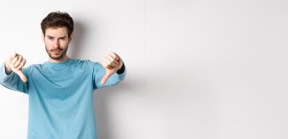 Skeptical handsome guy with beard, showing thumbs down in disapproval, dislike something bad, standing over white background photo