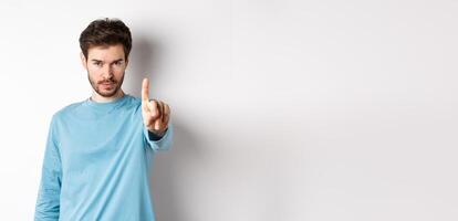 Image of serious young man with beard, shaking finger in disapproval, prohibit or forbid something, standing over white background and saying no photo