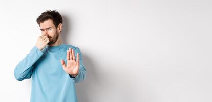 Image of caucasian man stretch out hands to stop or reject something, shut nose disgust, refusing product with bad smell, standing over white background photo