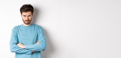 Image of offended and sad young man with beard, look from under forehead and sulking irritated, cross arms on chest defensive, mad at someone, standing on white background photo