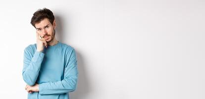 Skeptical young man listening to you with bored face, looking at camera reluctant, standing in sweatshirt over white background photo