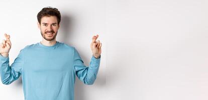 Optimistic smiling man cross fingers for good luck, making wish and waiting for results, standing over white background photo