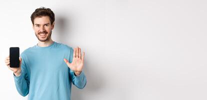 Handsome caucasian man with beard, showing empty smartphone screen and number five, raising hand to wave and say hello, standing on white background photo