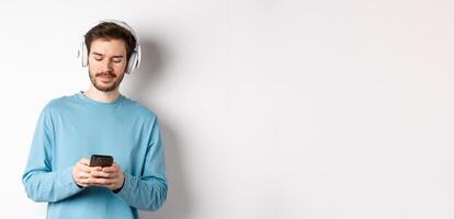 Modern young man in blue sweatshirt put on music on smartphone, listening songs in wireless headphones, white background photo