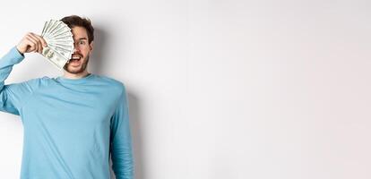Surprised and happy young man covering half of face with money, gasping amazed at camera, checking out quick loan offers, standing over white background photo
