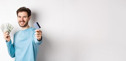 Smiling caucasian man holding money and giving you plastic credit card, standing on white background photo