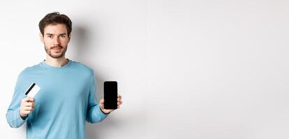 Handsome caucasian man showing empty smartphone screen and plastic credit card, standing over white background photo