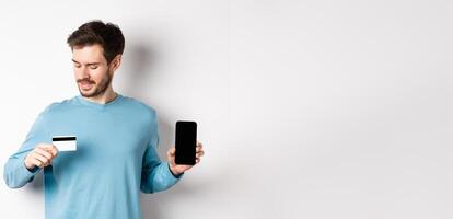 Handsome young man with beard, showing empty smartphone screen and looking at plastic credit card, standing over white background photo