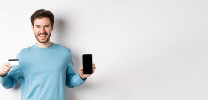 Smiling caucasian man showing plastic credit card with mobile phone screen. Guy recommending online banking app, standing on white background photo