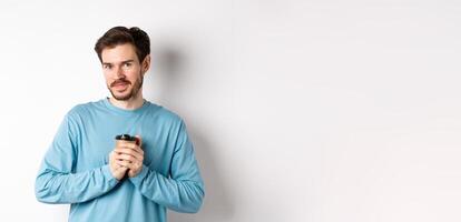 Handsome caucasian man warm-up hands with cup of coffee, smiling at camera and looking tenderly, standing on white background photo
