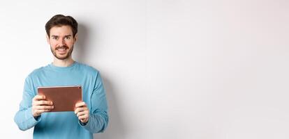 Handsome young man holding digital tablet and smiling, networking with device, standing over white background photo