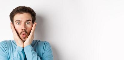 Close-up of impressed man checking out special offer, saying wow and touching face with amazed expression, stare at camera in awe, standing over white background photo
