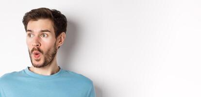 Close-up of impressed caucasian man saying wow, looking left amazed, checking out promotion deal, white background photo