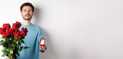 Valentines and relationship. Romantic man boyfriend holding red roses and showing engagement ring, making a proposal on lovers day, standing over white background photo